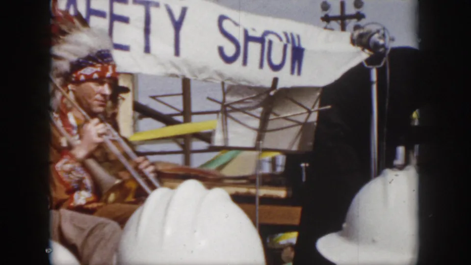 1958 Virginia 8mm frame showing a safety show performer at left, white hard hats in the foreground, and a partial suited figure crossing the stage area.
