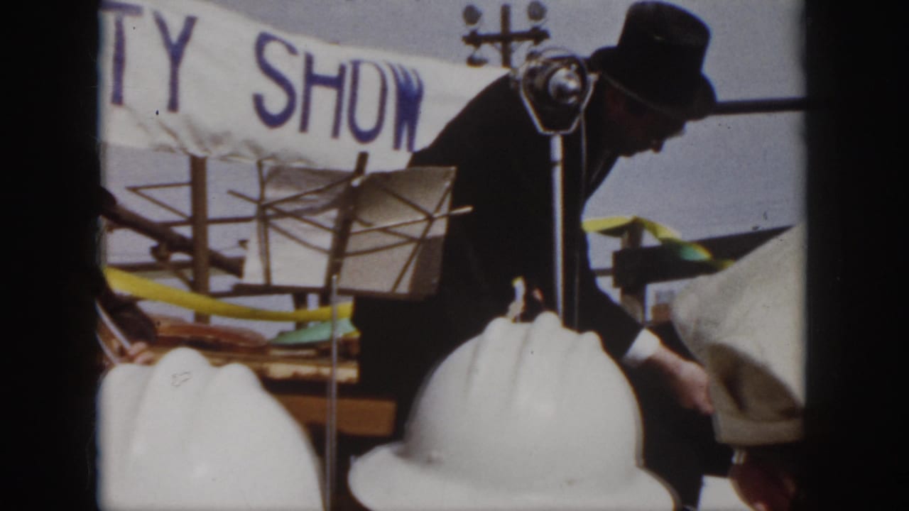 1958 Virginia workplace safety show frame with a dark-suited man bending near the stage, white hard hats below, and the banner behind him.