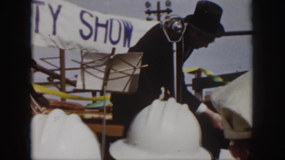 1958 Virginia workplace safety show frame with a dark-suited man bending near the stage, white hard hats below, and the banner behind him.