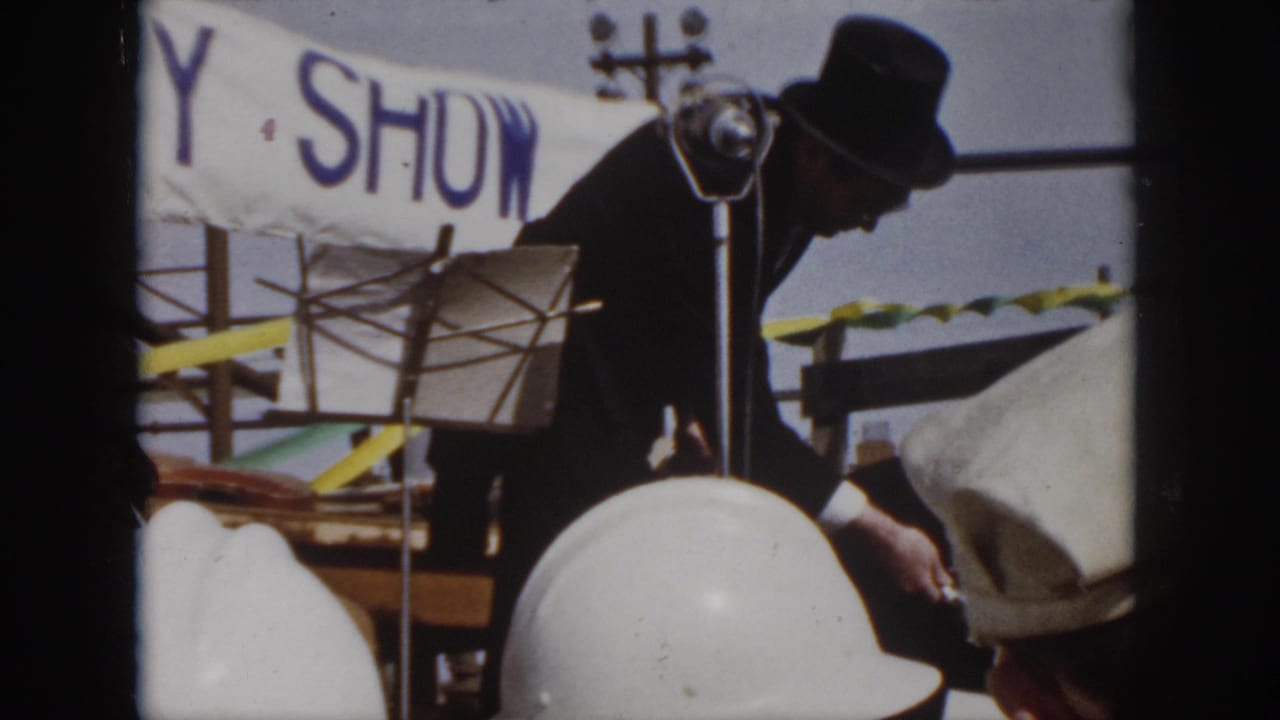 1958 Virginia 8mm frame showing two magicians in dark coats and top hats on an outdoor stage with microphones and fluttering streamers.