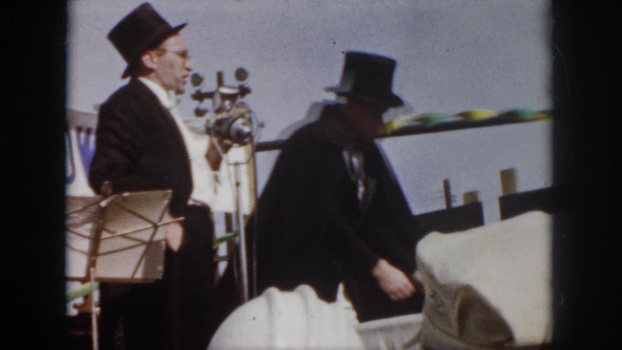 A top-hatted magician stands at a microphone while another performer moves at the side of the stage.