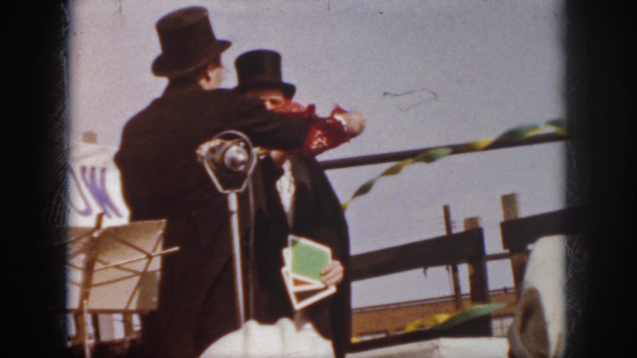 1958 Virginia frame showing two magicians in top hats close together on an outdoor stage, with a red cloth prop between them.