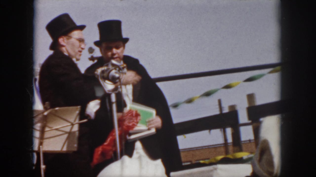 1958 Virginia home-movie frame showing a man in a top hat at a microphone while another man stands beside him.