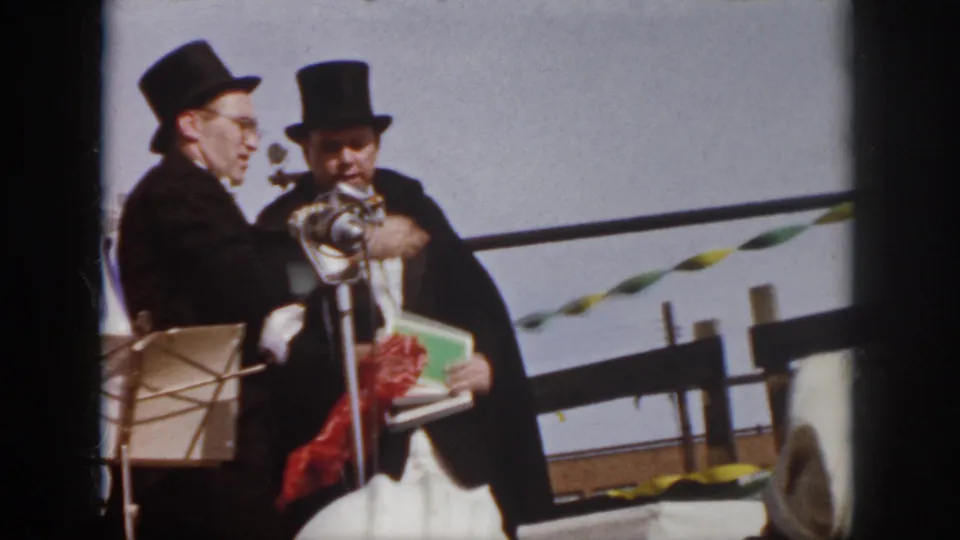 1958 Virginia home-movie frame showing a man in a top hat at a microphone while another man stands beside him.