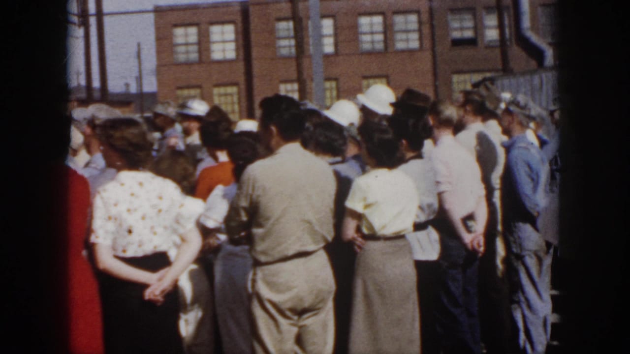 1958 Virginia home-movie view of a standing crowd in mid-century clothing watching an outdoor event near a brick building.