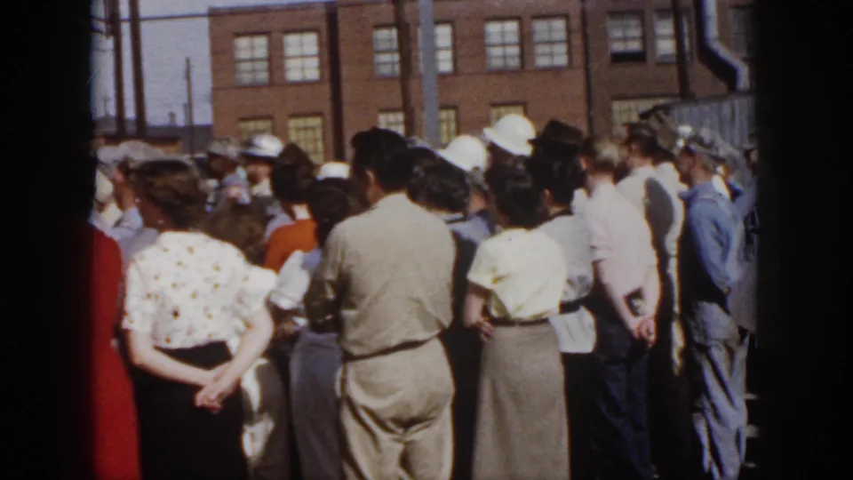 1958 Virginia home-movie view of a standing crowd in mid-century clothing watching an outdoor event near a brick building.