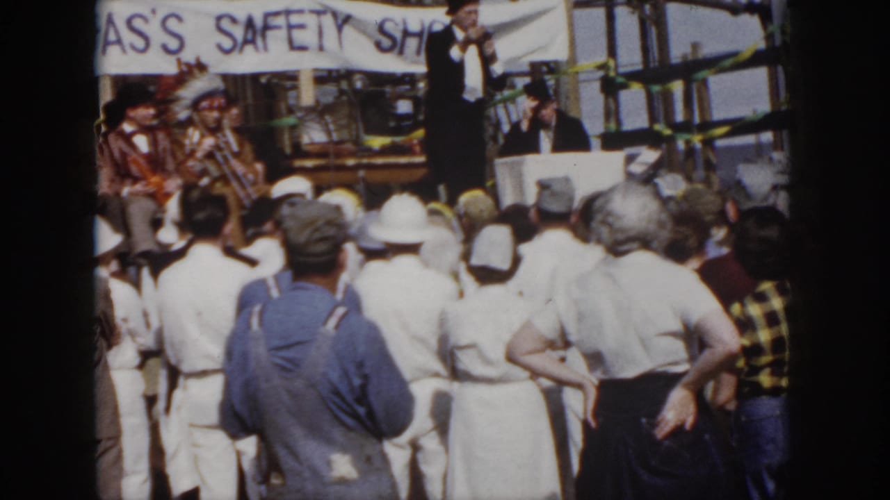 1958 Virginia 8mm frame of workers in hard hats and uniforms facing a suited speaker on an outdoor industrial stage.