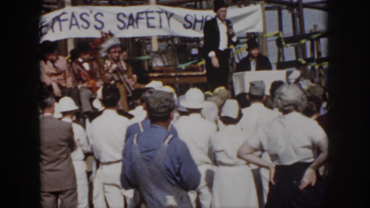 Workers stand in front of a stage with a speaker and seated performers under a banner.
