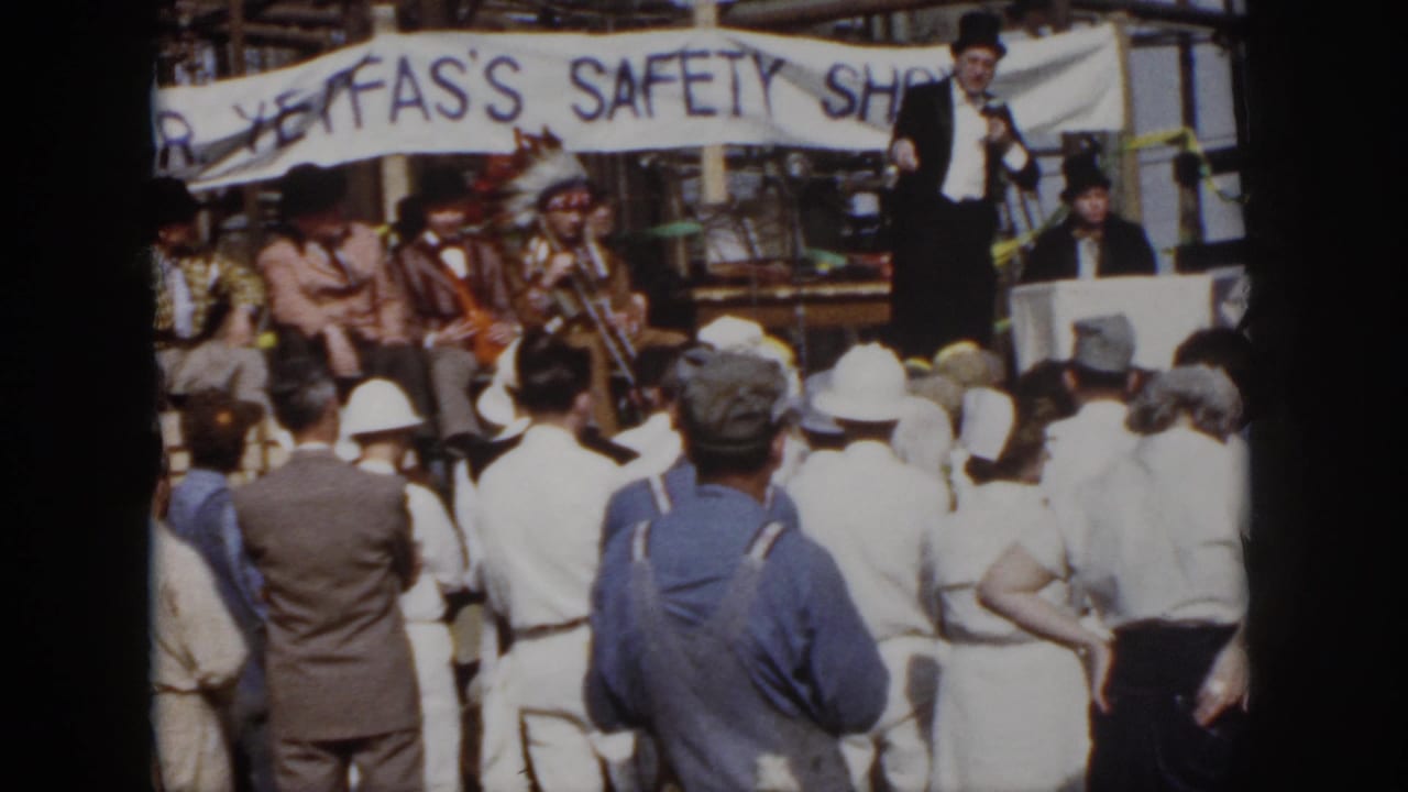 1958 Virginia 8mm view of an industrial crowd watching a microphone speaker with seated musicians beneath a safety banner.