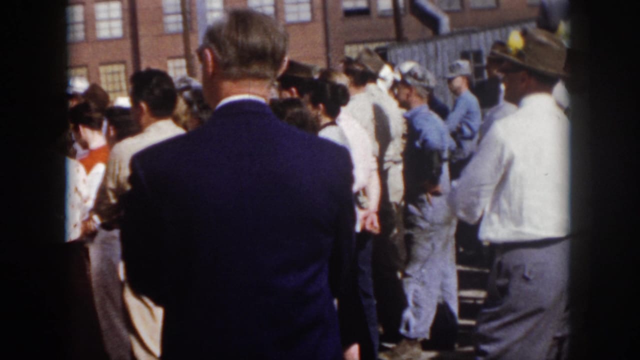 1958 Virginia 8mm frame looking through the crowd at workers and attendees near brick industrial buildings after the stage view shifts.