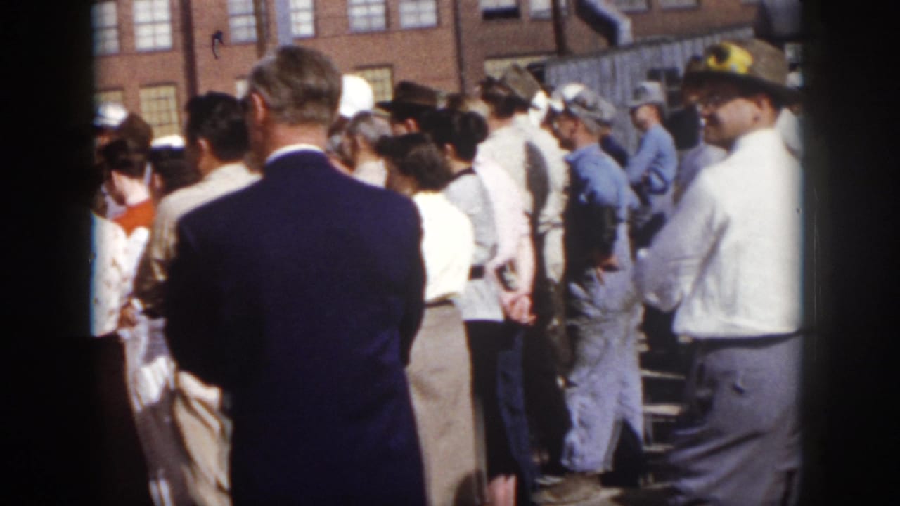 Adults gather outside a brick industrial building, with a suited man seen from behind and workers nearby.