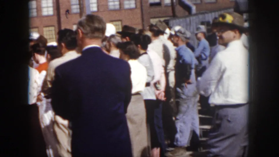 1958 Virginia 8mm frame showing adults outside a brick industrial building, including a suited man, hats, hard hats and blue work clothes.