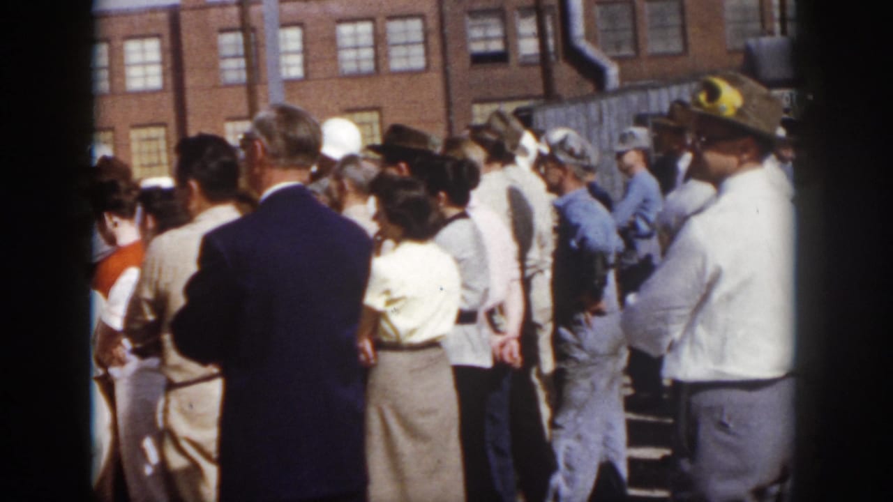 1958 Virginia 8mm frame of adults gathered outdoors at an industrial building, mixing civilian clothing with work coveralls and hard hats.