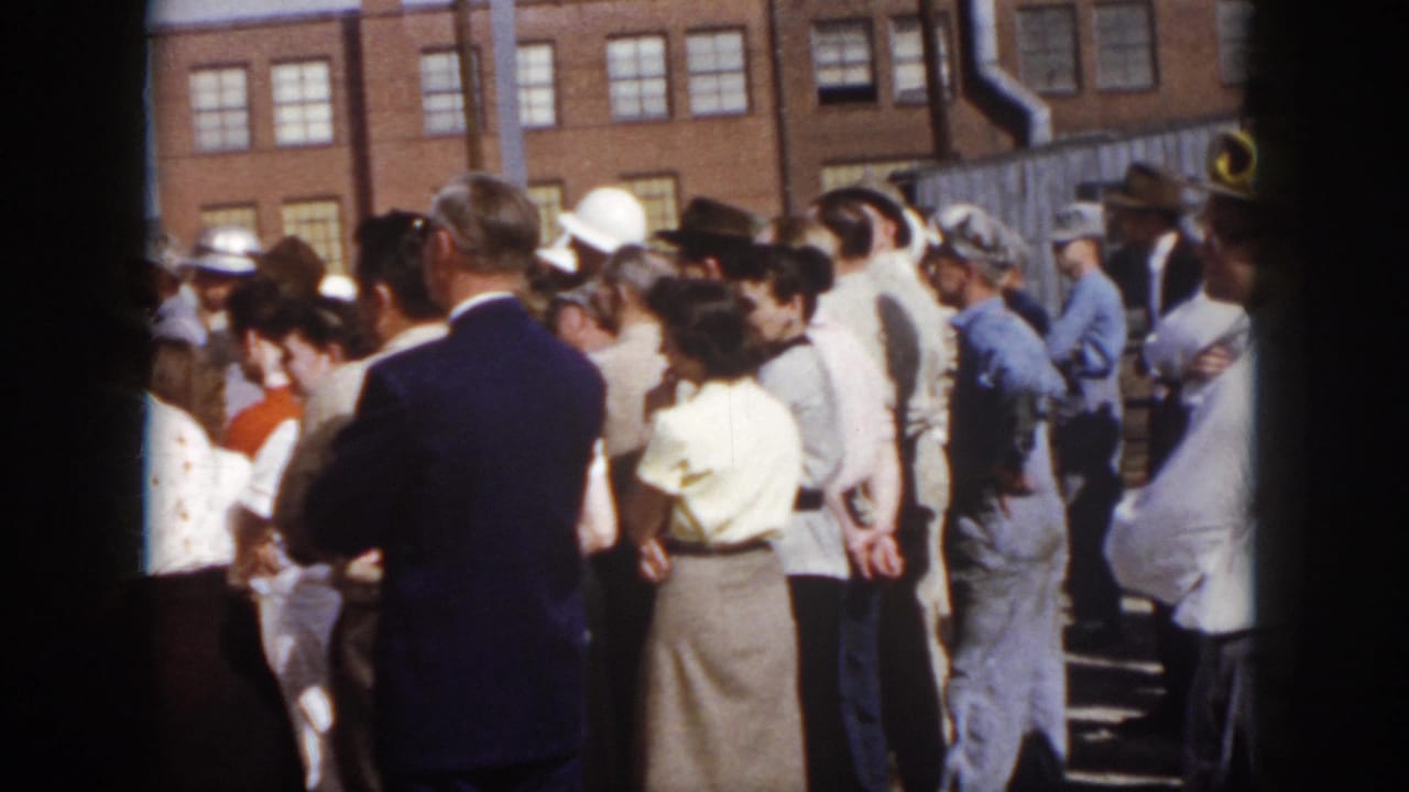 1958 Virginia 8mm frame showing a gathered adult crowd from behind, with period clothing and industrial workers beside a brick exterior.