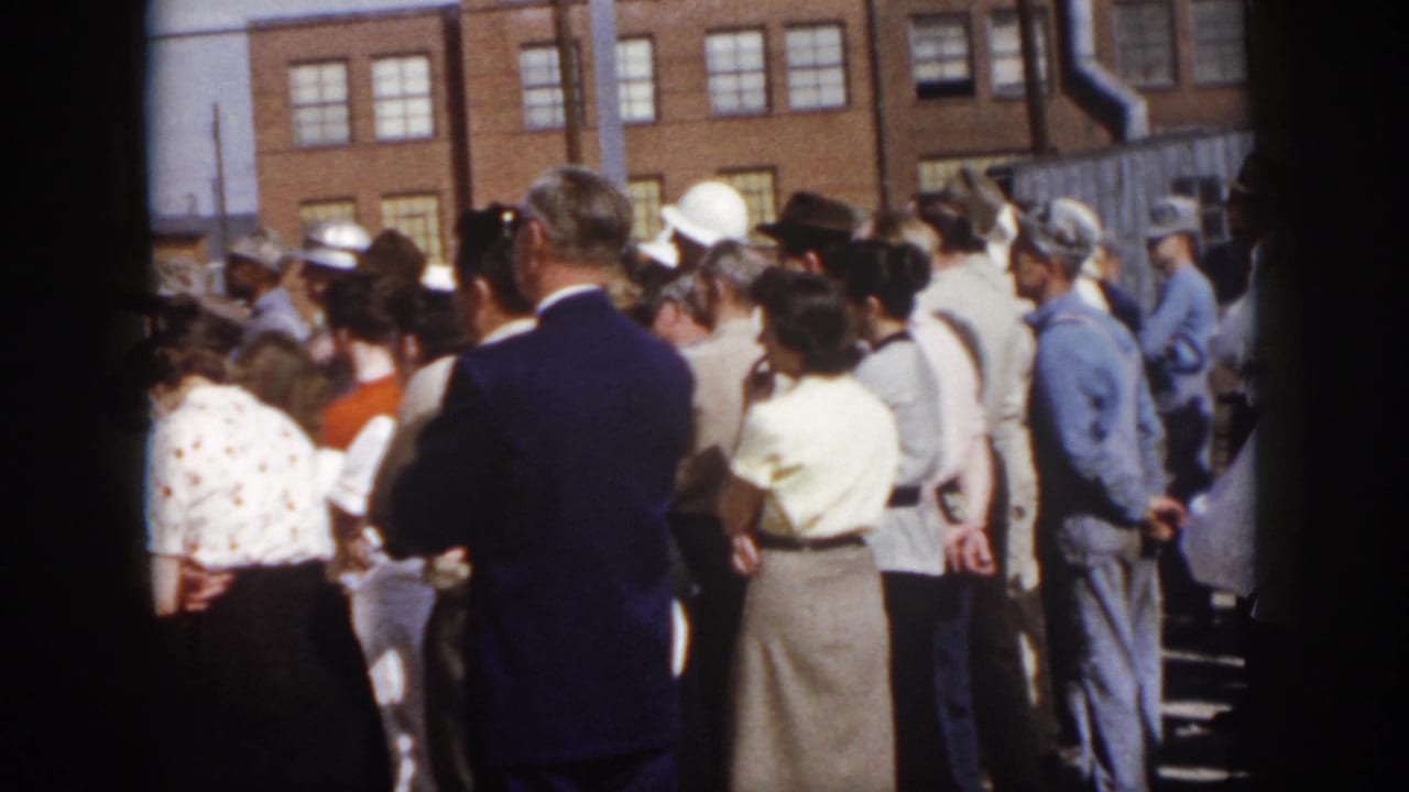1958 Virginia 8mm frame of a mixed adult crowd outside a brick industrial building, including suited civilians and workers in blue coveralls.