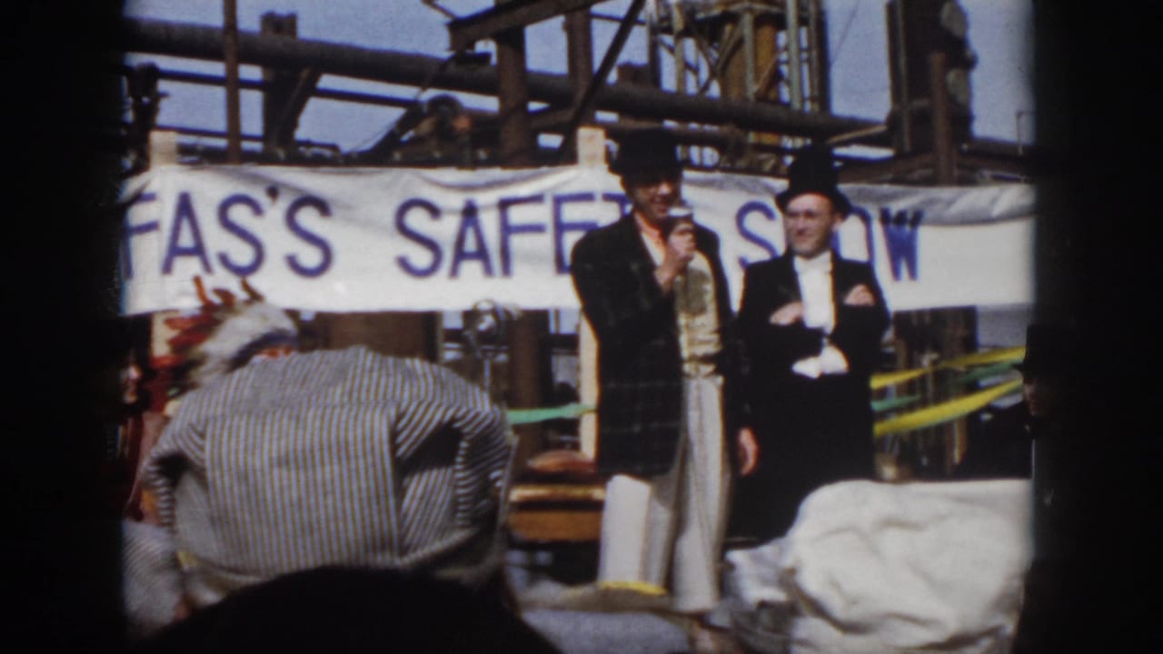 Outdoor 1958 Virginia safety show frame with two formally dressed men, a microphone, banner text, and audience foreground.