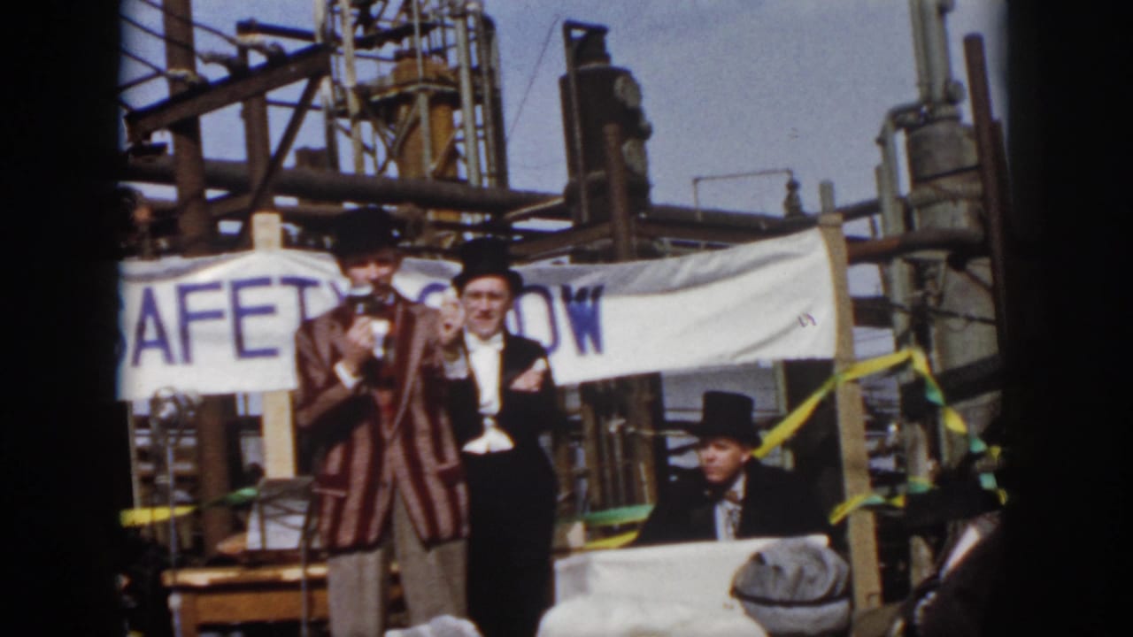 Virginia 1958 home-movie frame showing costumed men with top hats on a small stage before a safety-show banner and industrial equipment.