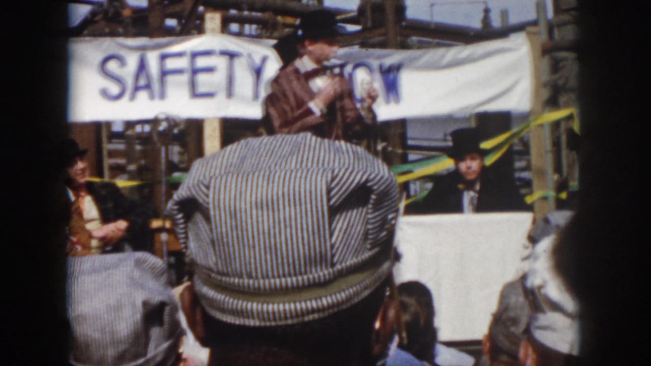 1958 Virginia home-movie frame showing attendees facing a stage area with a SAFETY SHOW banner and industrial structures behind it.