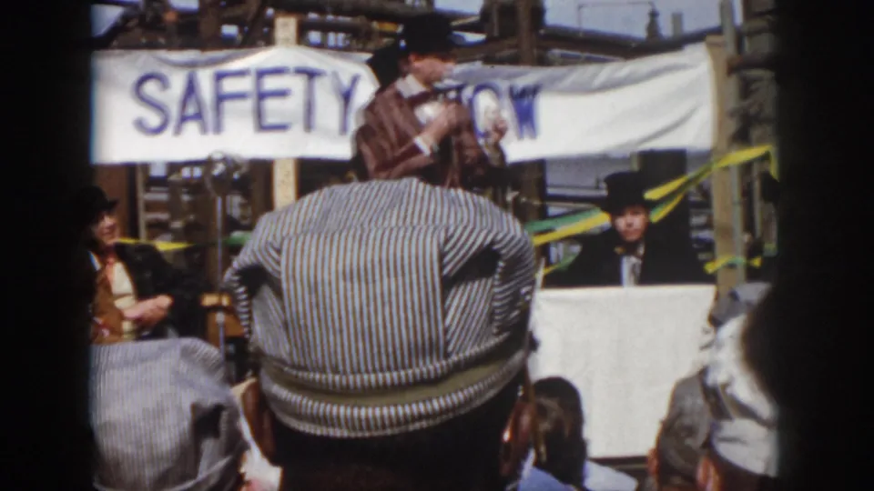 1958 Virginia home-movie frame showing attendees facing a stage area with a SAFETY SHOW banner and industrial structures behind it.