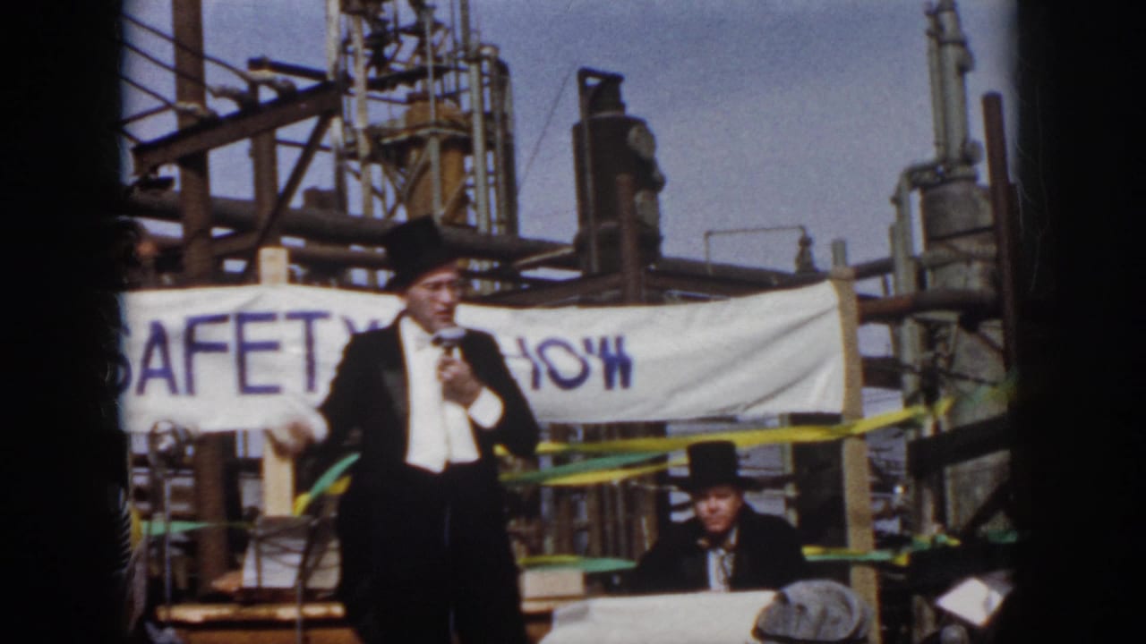 Home-movie frame from the Virginia industrial safety event showing the speaker centered against a banner, pipes, tanks, and a seated assistant.