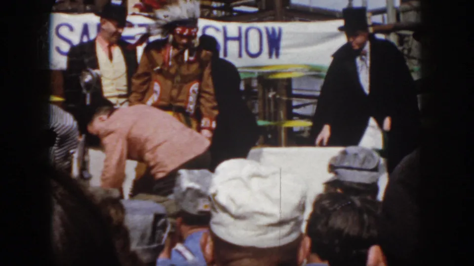 1958 Virginia safety-show scene with a man in Native American regalia, a tuxedoed figure, audience members, and industrial backdrop.