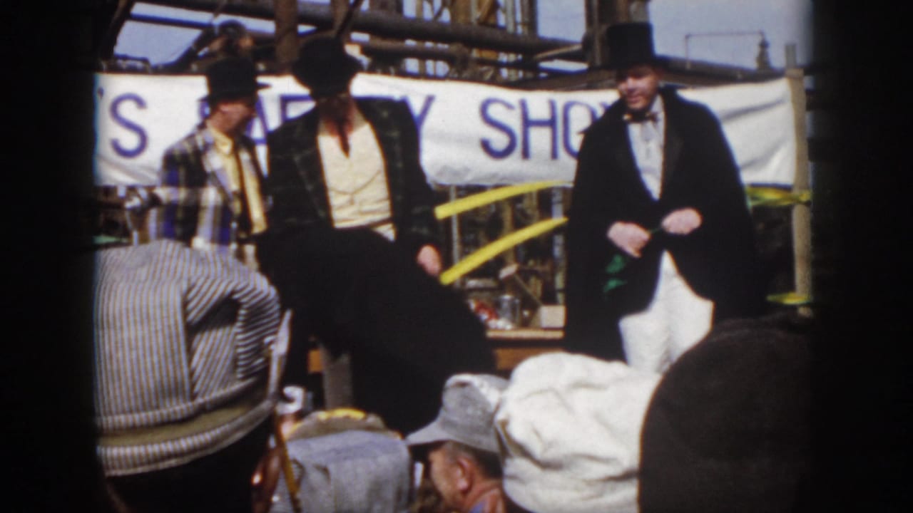 A caped performer and other costumed men stand before seated spectators.