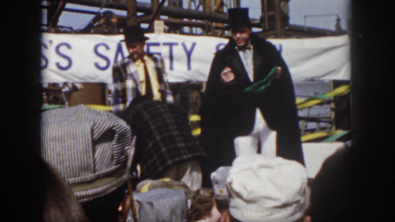 1958 Virginia frame with a top-hatted man holding a green prop as another performer bends near the audience during the dockside safety show.