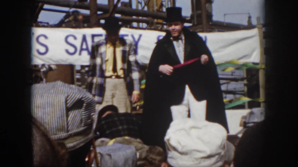 1958 Virginia home-movie frame showing a magician on an outdoor stage with an assistant, seated spectators, and a visible safety banner behind them.