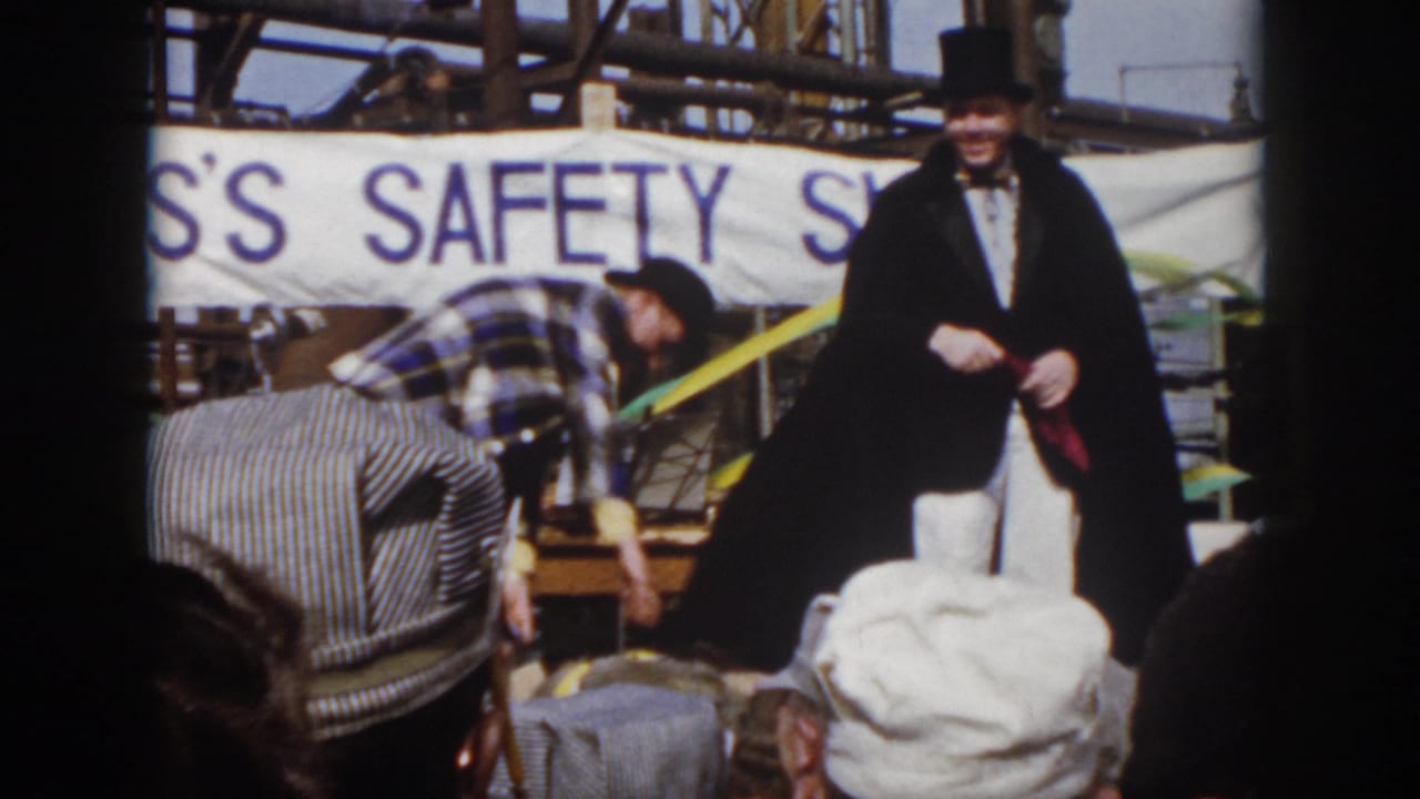 1958 Virginia outdoor event frame with the magician standing near the edge of the stage while the assistant reaches down by props and the crowd watches.