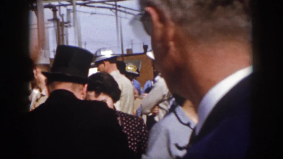 1958 Virginia event frame from behind the audience, showing the magician's top hat, nearby spectators, and uniformed or work-clothed people near the stage area.