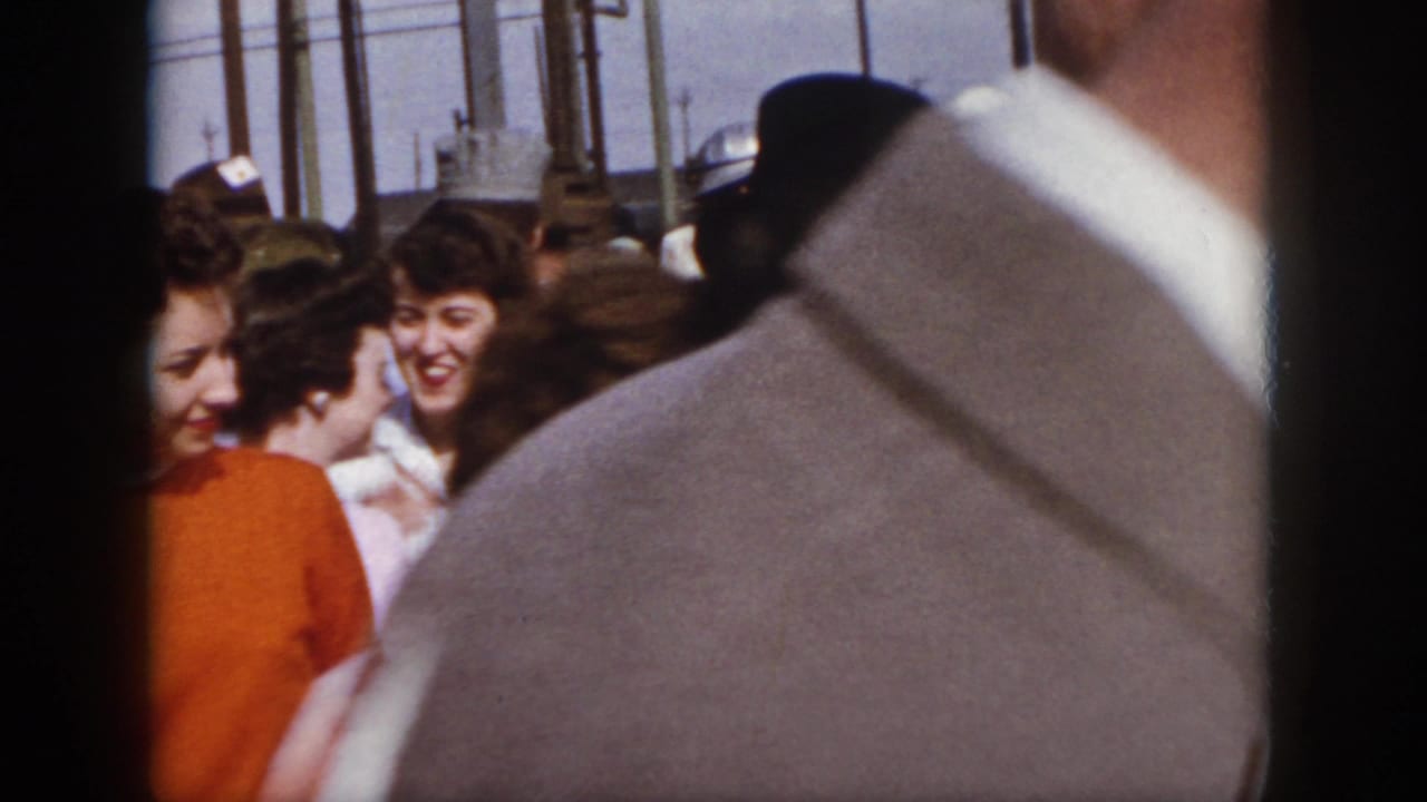 1958 Virginia home-movie frame showing a crowded outdoor walkway, partly obscured by a man in a gray jacket with women moving behind him.