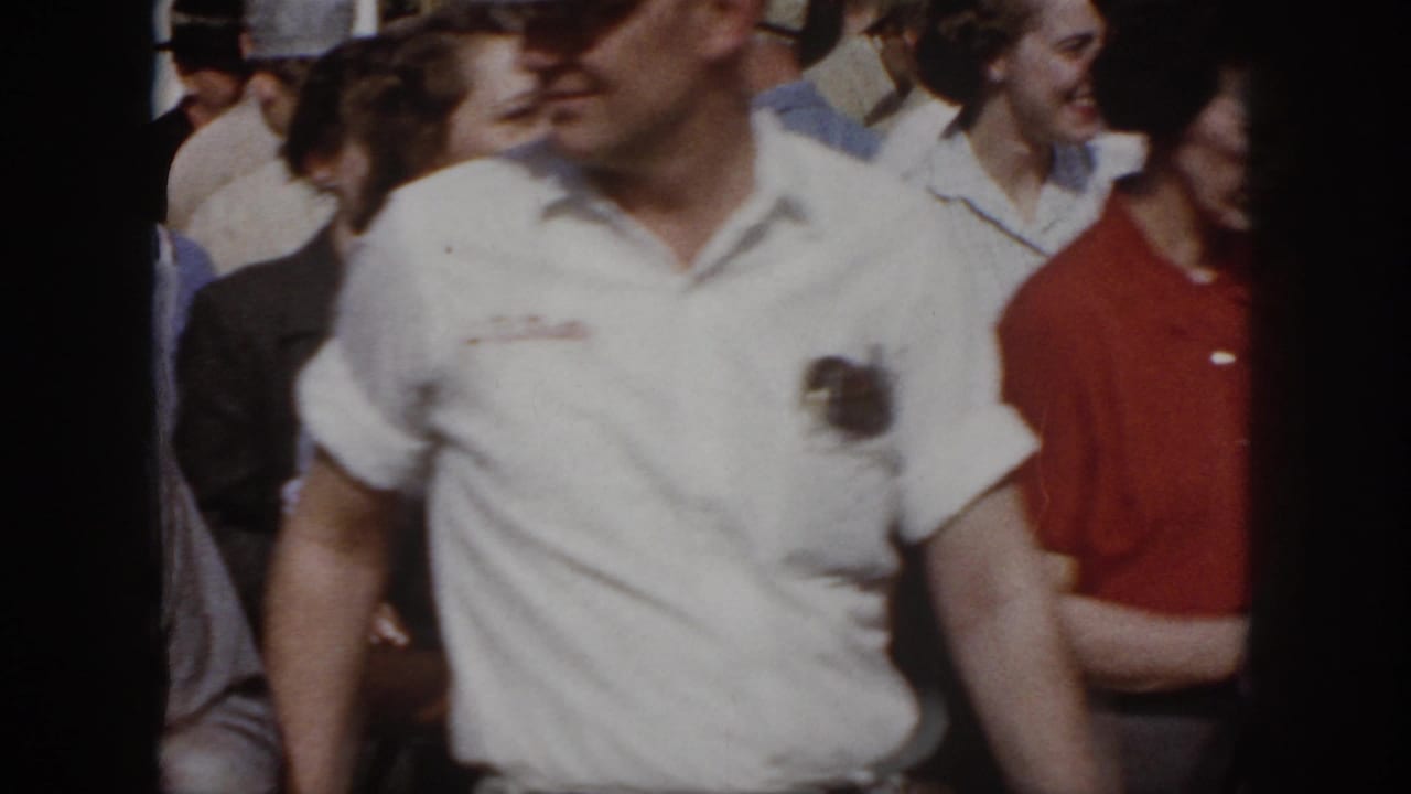 1958 Virginia frame with a man in a white uniform-style shirt crossing close to the camera as adults move behind him.