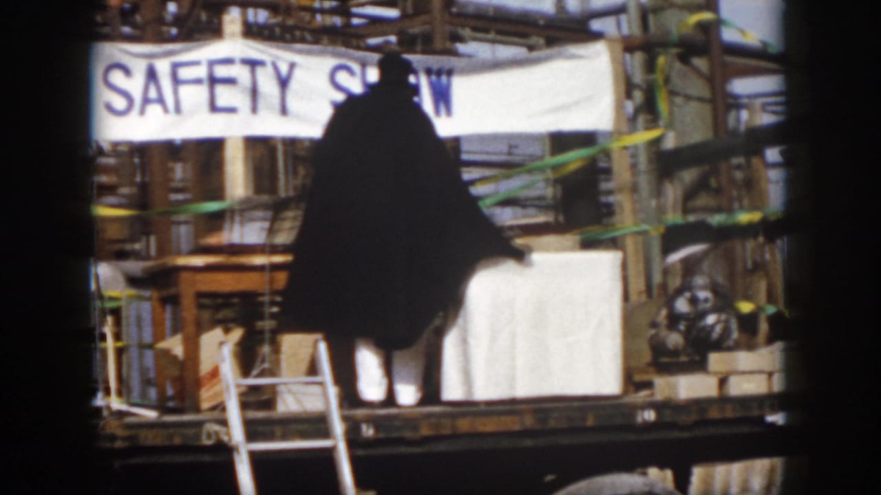 Caped figure on a wooden platform near a SAFETY SHOW banner and ladders.