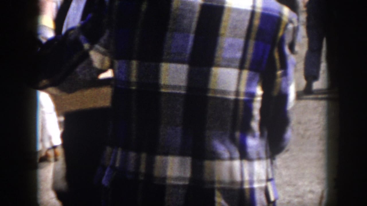 A handheld 8mm view follows fairgoers walking near the safety show area, with a blue plaid shirt prominent in the foreground.
