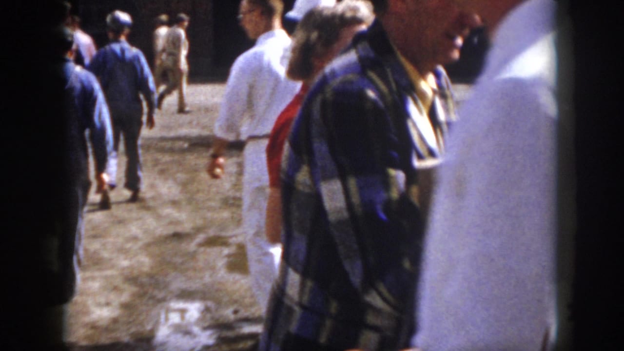 1958 Virginia home-movie frame showing men in an industrial yard, with brick buildings and foreground figures partly blocking the view.