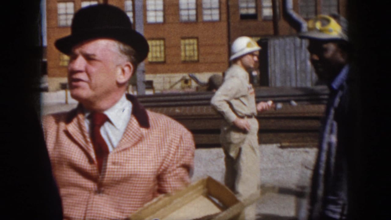 Man in a bowler hat and checkered jacket stands near hard-hatted workers and stacked rails.