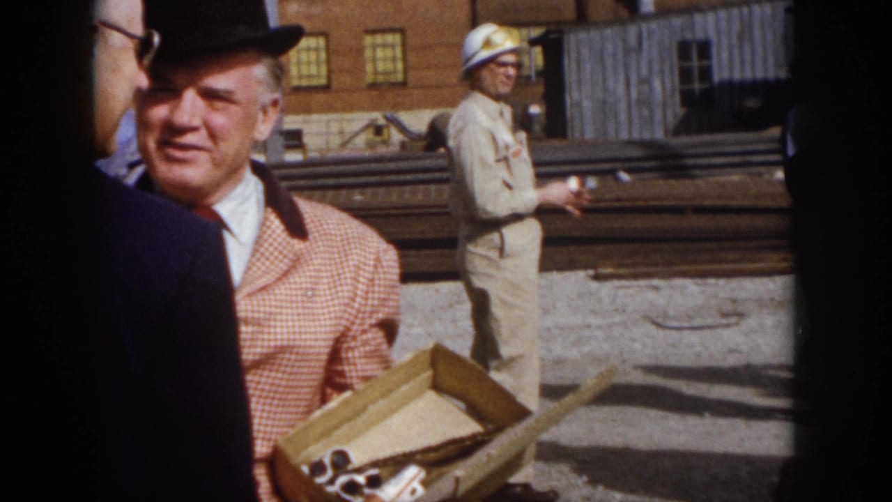 1958 Virginia home-movie frame of a bowler-hatted man beside a box, with a hard-hatted worker and stacked rails behind him.