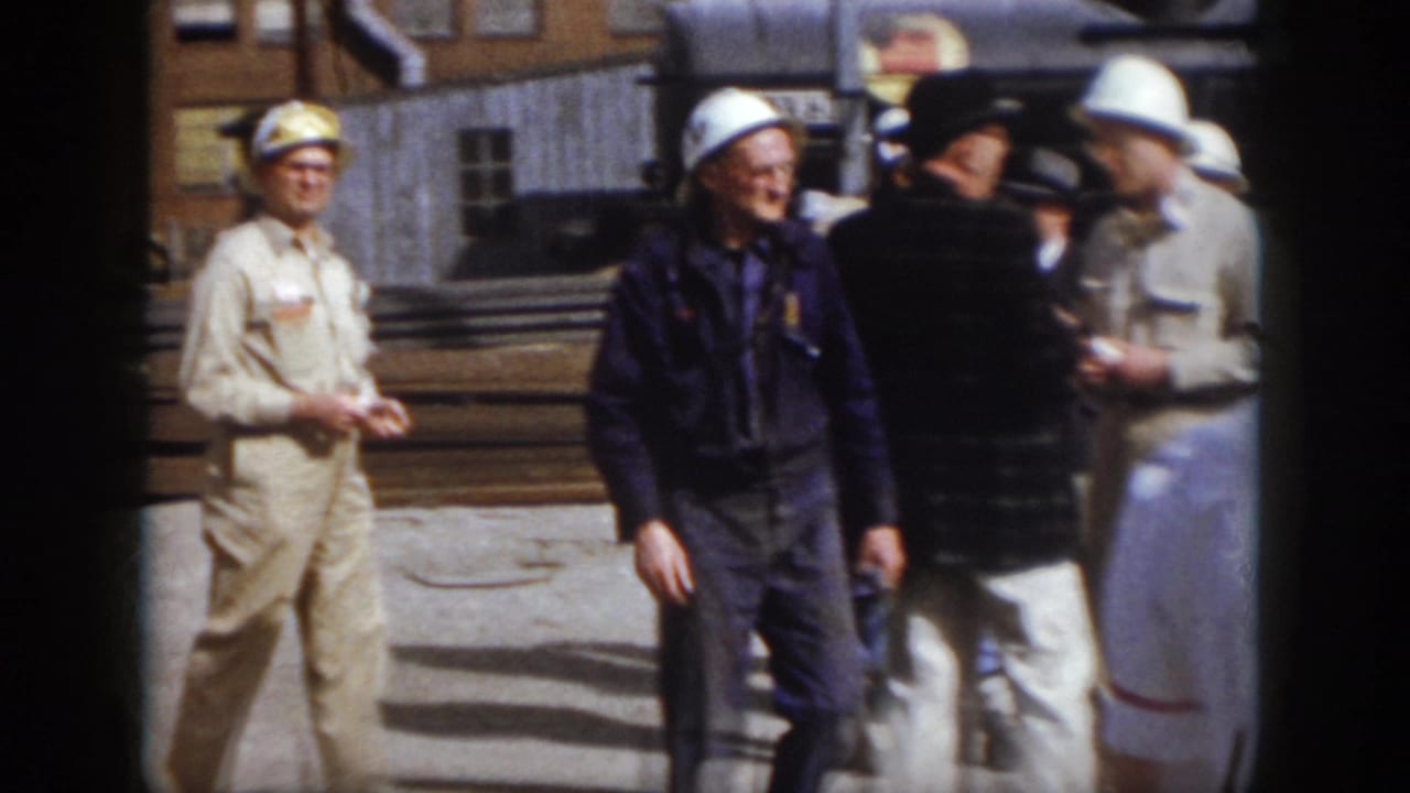 1958 Virginia industrial yard frame showing hard-hatted workers and men in work clothes gathered near stacked rails and buildings.