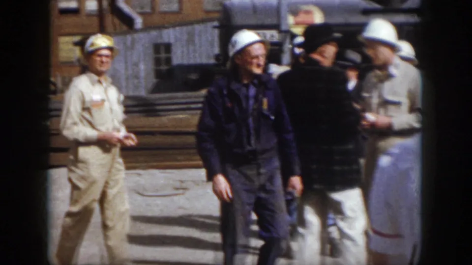 1958 Virginia industrial yard frame showing hard-hatted workers and men in work clothes gathered near stacked rails and buildings.