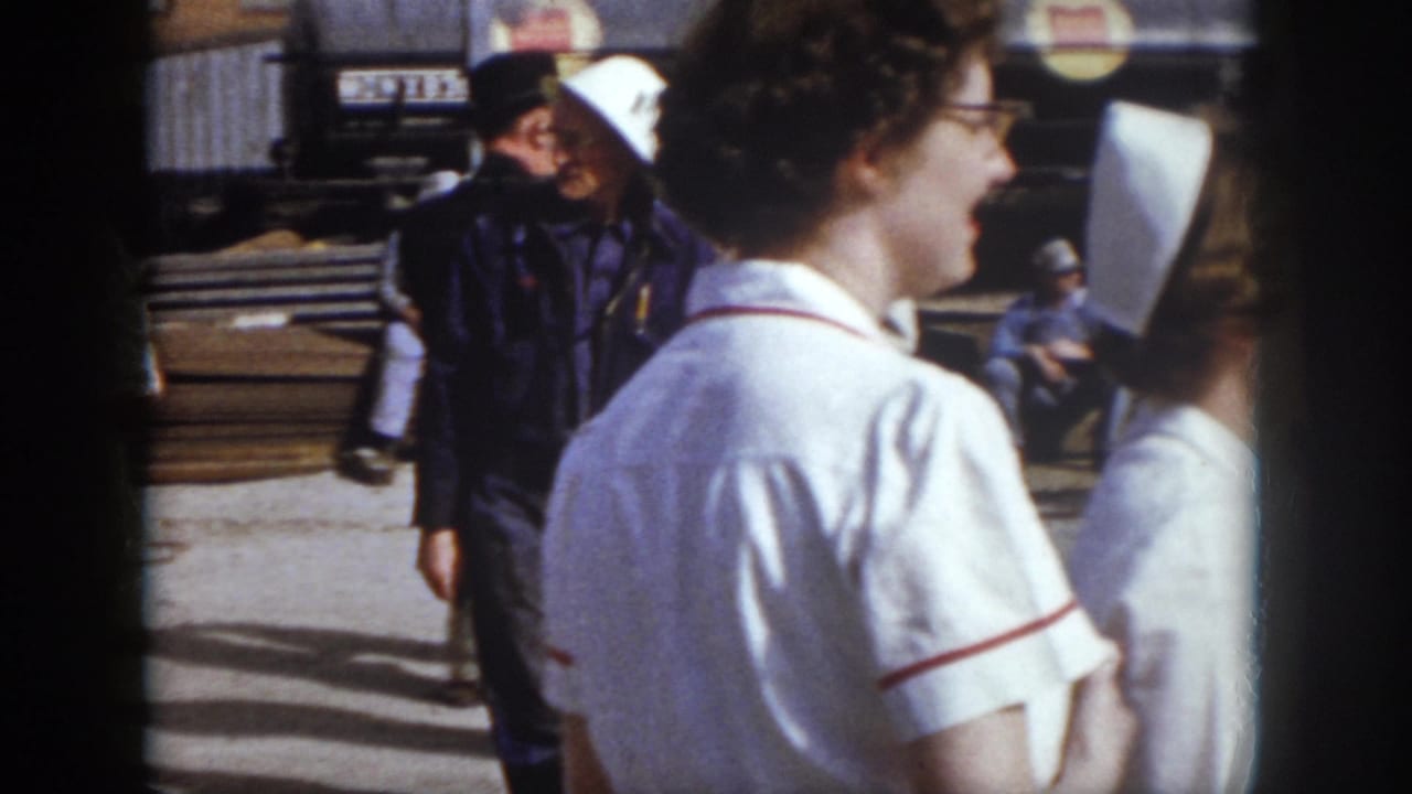 1958 Virginia home-movie frame showing nurses in white caps near men in workwear and hard hats on a concrete pad.