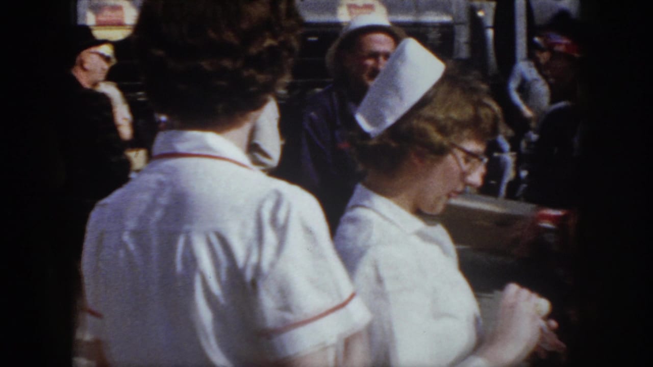 Two nurses in white uniforms stand near workers and spectators outdoors.