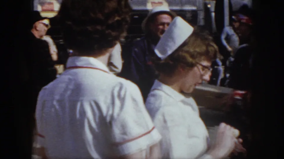 1958 Virginia frame with nurses in white caps beside men in hard hats and other adults in an outdoor gathering.