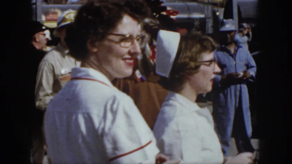1958 Virginia home-movie still of a smiling nurse with glasses beside another uniformed woman at an outdoor gathering.