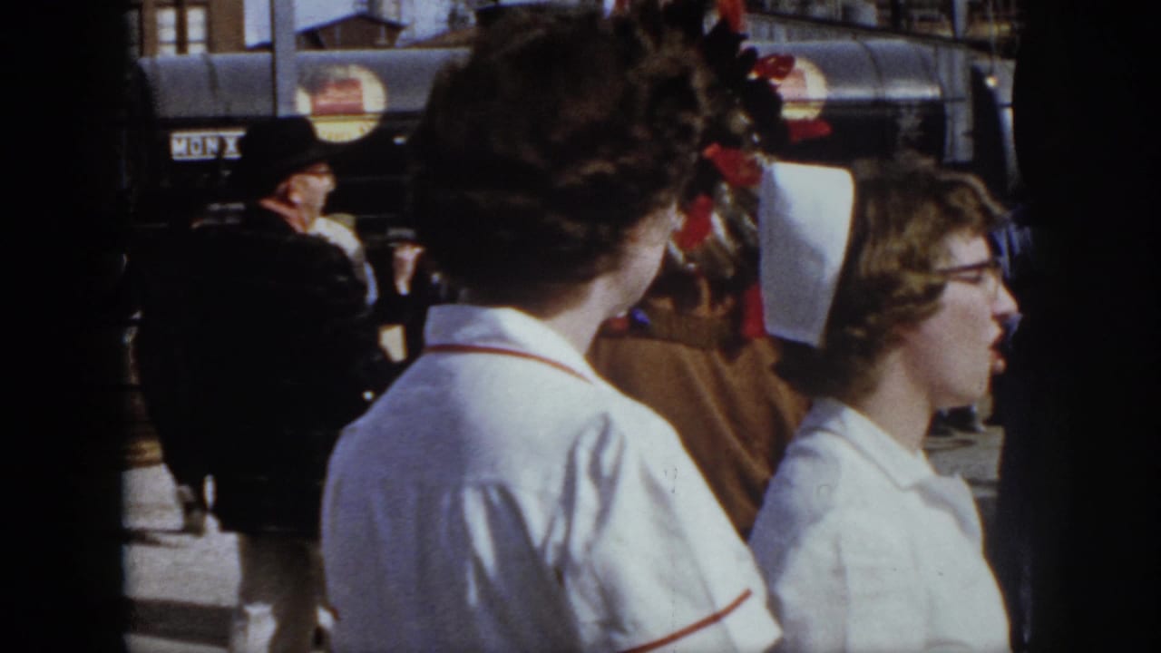 1958 Virginia frame showing nurses in white caps near workers and a person with feathered decoration in the background.
