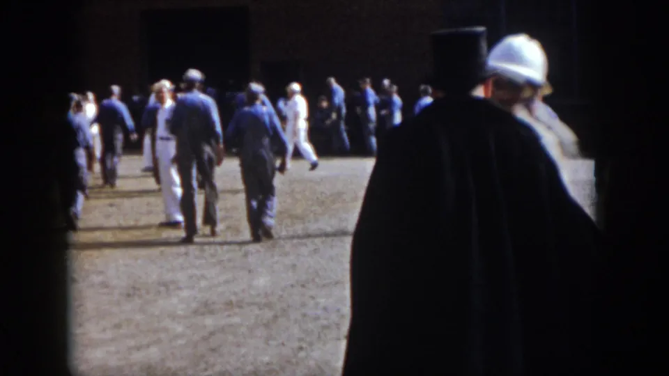 1958 Virginia home-movie frame showing men walking and milling outdoors, with dark foreground obstruction and bright pavement.