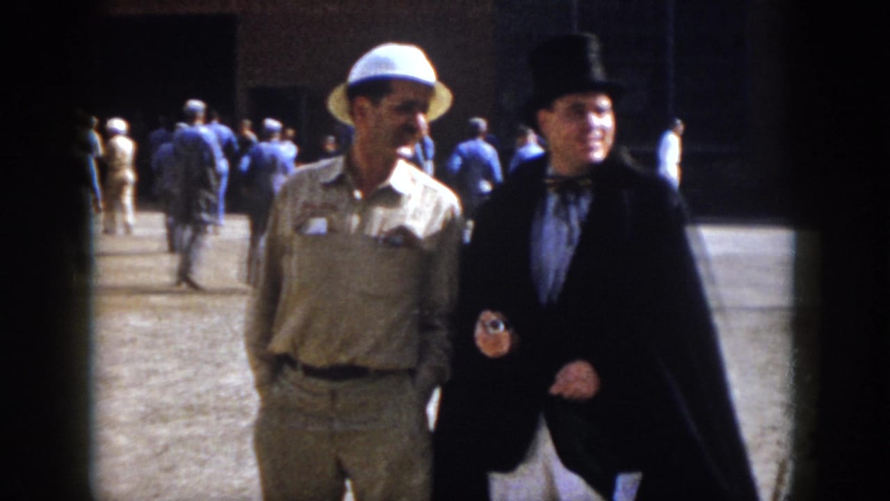 1958 Virginia home-movie frame showing two men walking together, one in safari-style clothing and one in a black top hat and cape.