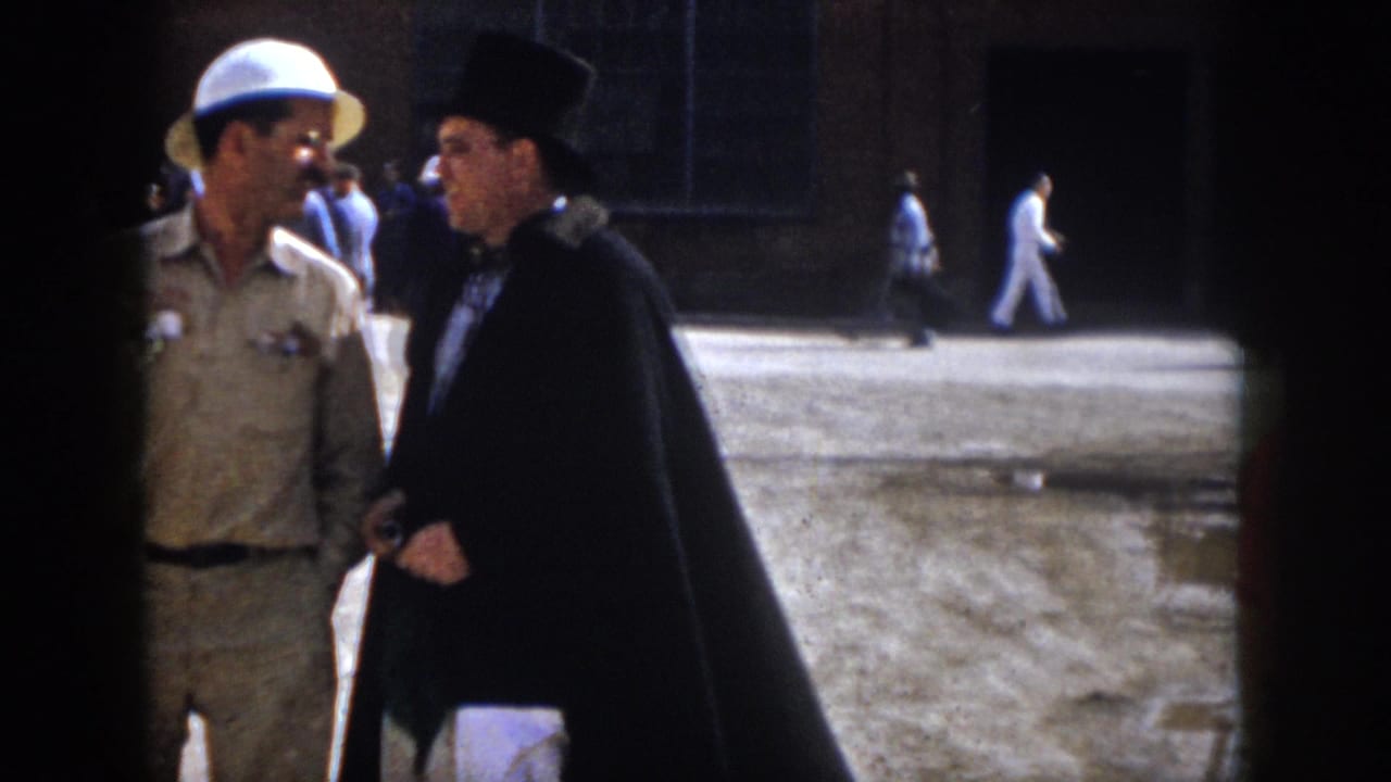1958 Virginia frame showing a close outdoor conversation between a khaki-clad man and a man in a black top hat and cape.