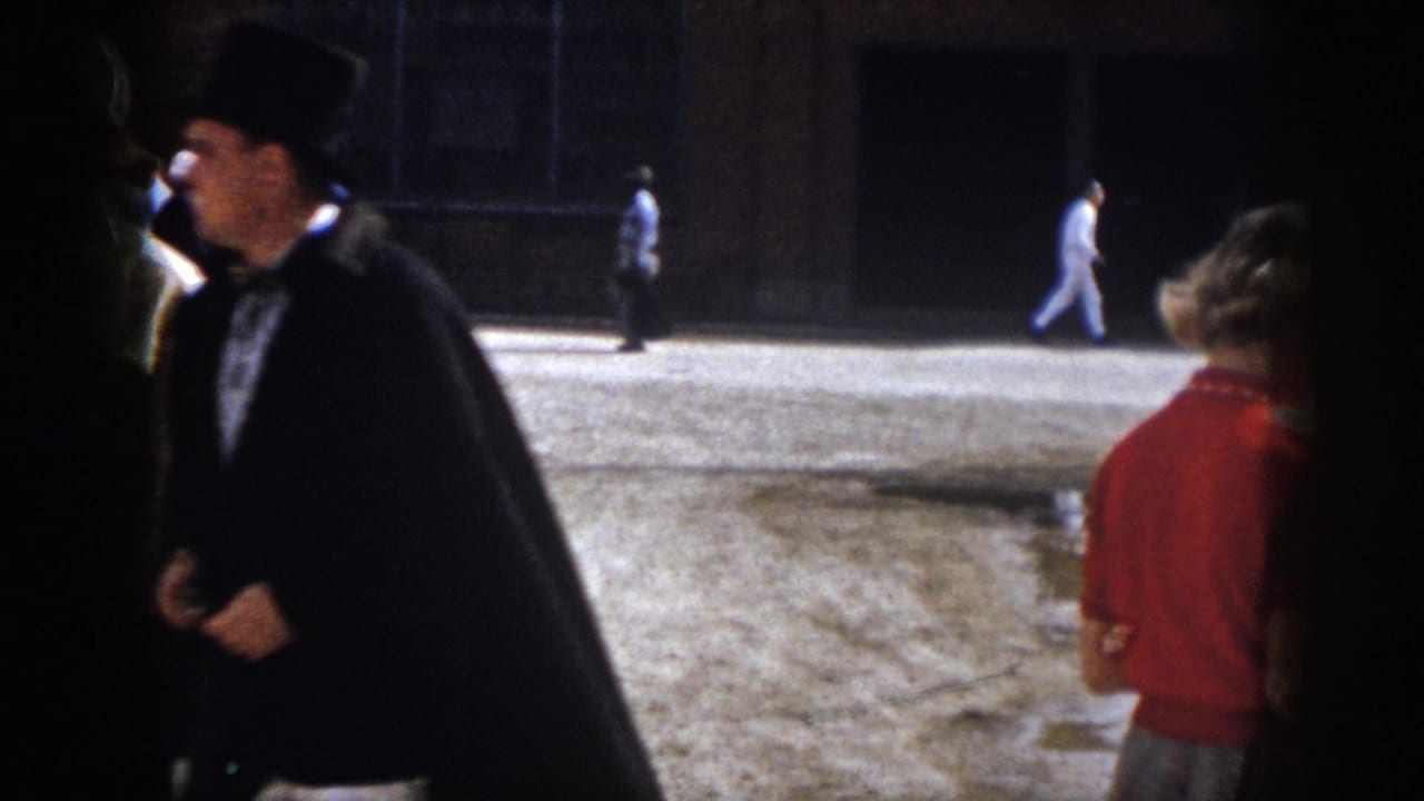 1958 Virginia home-movie frame showing a caped man in a top hat beside a woman in red as pedestrians cross a dirt lot.
