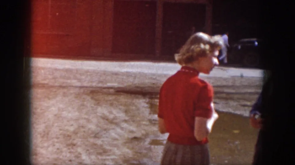 1958 Virginia frame with a woman in a red blouse and skirt standing near a muddy lot and building frontage.