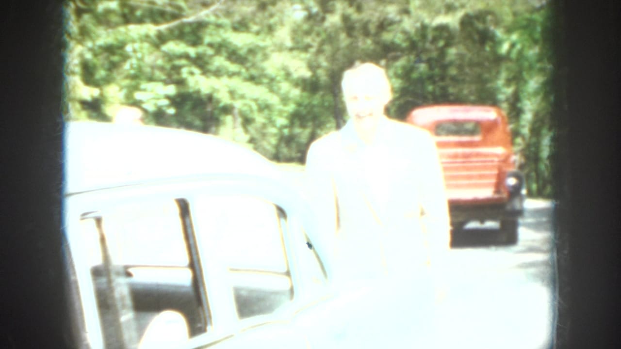 Washed-out 1958 Virginia home-movie frame showing a person beside a light-colored car with a red truck farther back.